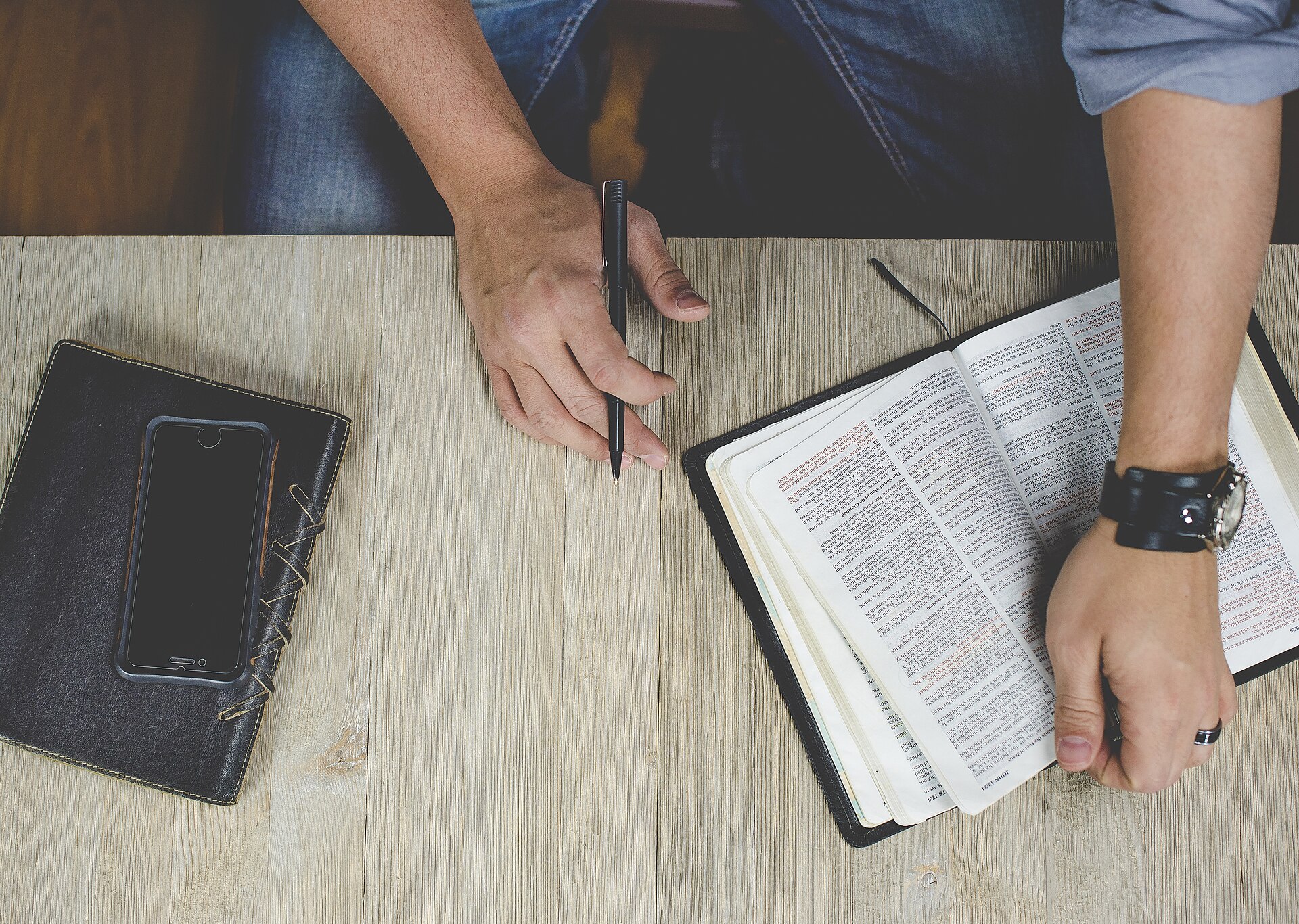 An open Bible and a notebook on a wooden desk, with a hand holding a pen
